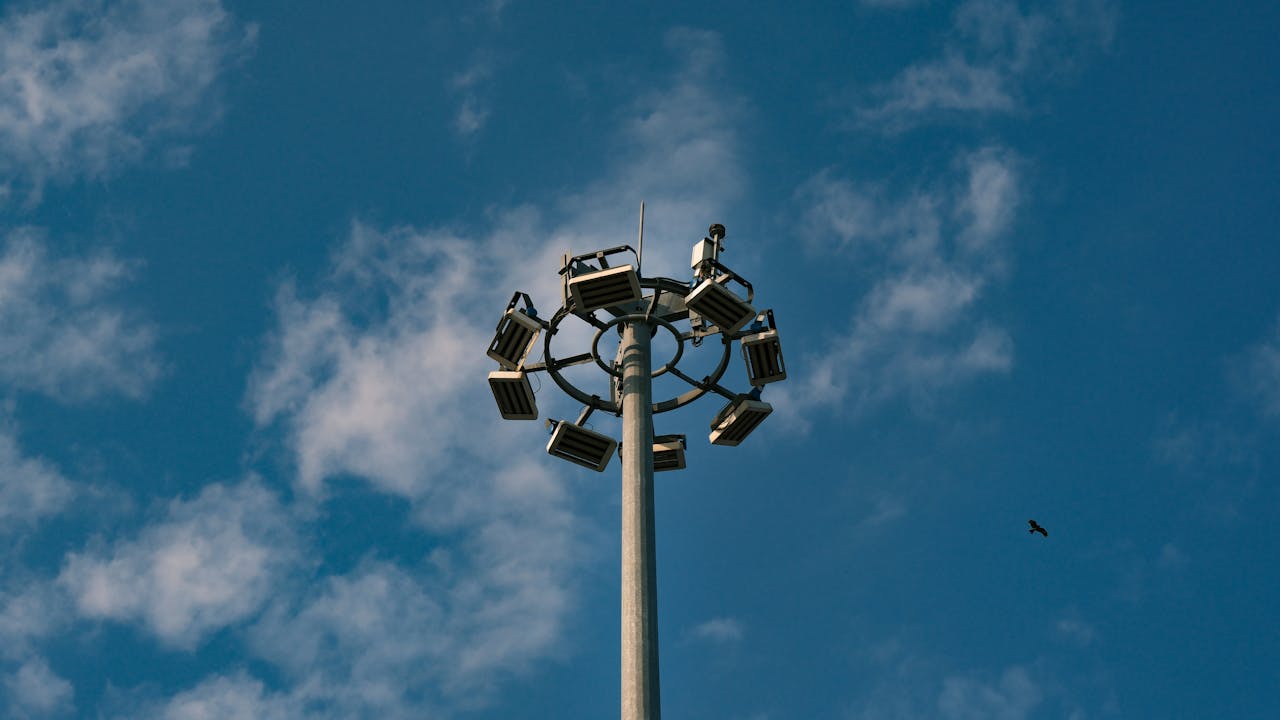 A tall lighting pole with multiple floodlights set against a bright blue sky with scattered clouds.